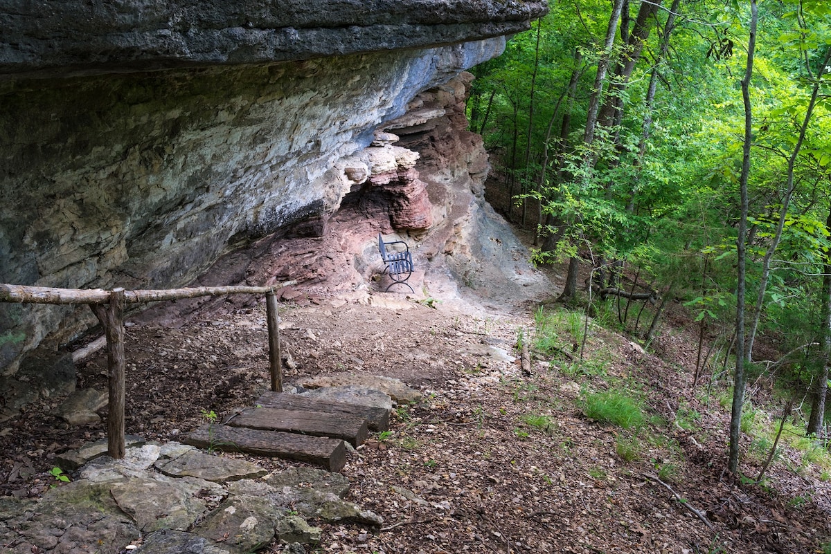 A natural rock formation is set against a lush green backdrop, with earth-toned hues visible on the cliff's surface. A wooden railing leads to a flat area containing scattered stones. Sunlight filters through the trees, creating dappled light on the forest floor.