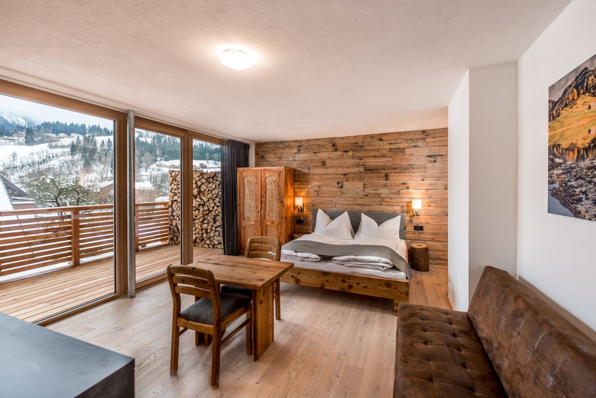 A cozy living area is complemented by a spacious bed and a wooden table with chairs. Large glass doors provide access to a balcony with scenic views of the outdoor landscape, while natural wood accents add warmth to the space.