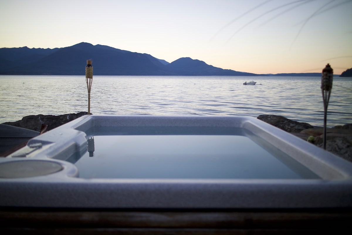 A hot tub is positioned on a wooden deck, overlooking calm waters that reflect the soft hues of sunset. Mountains are visible in the distance, with a small boat appearing on the water's surface. Tiki torches flank the scene, adding to the tranquil ambiance.