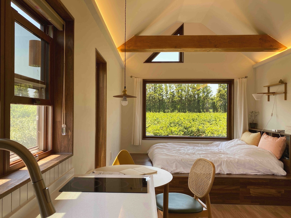 A bright interior space showcases a cozy sleeping area with a spacious bed. Natural light flows through large windows that frame views of lush greenery. A dining table and kitchen area are visible, complemented by wooden beams and stylish decor.