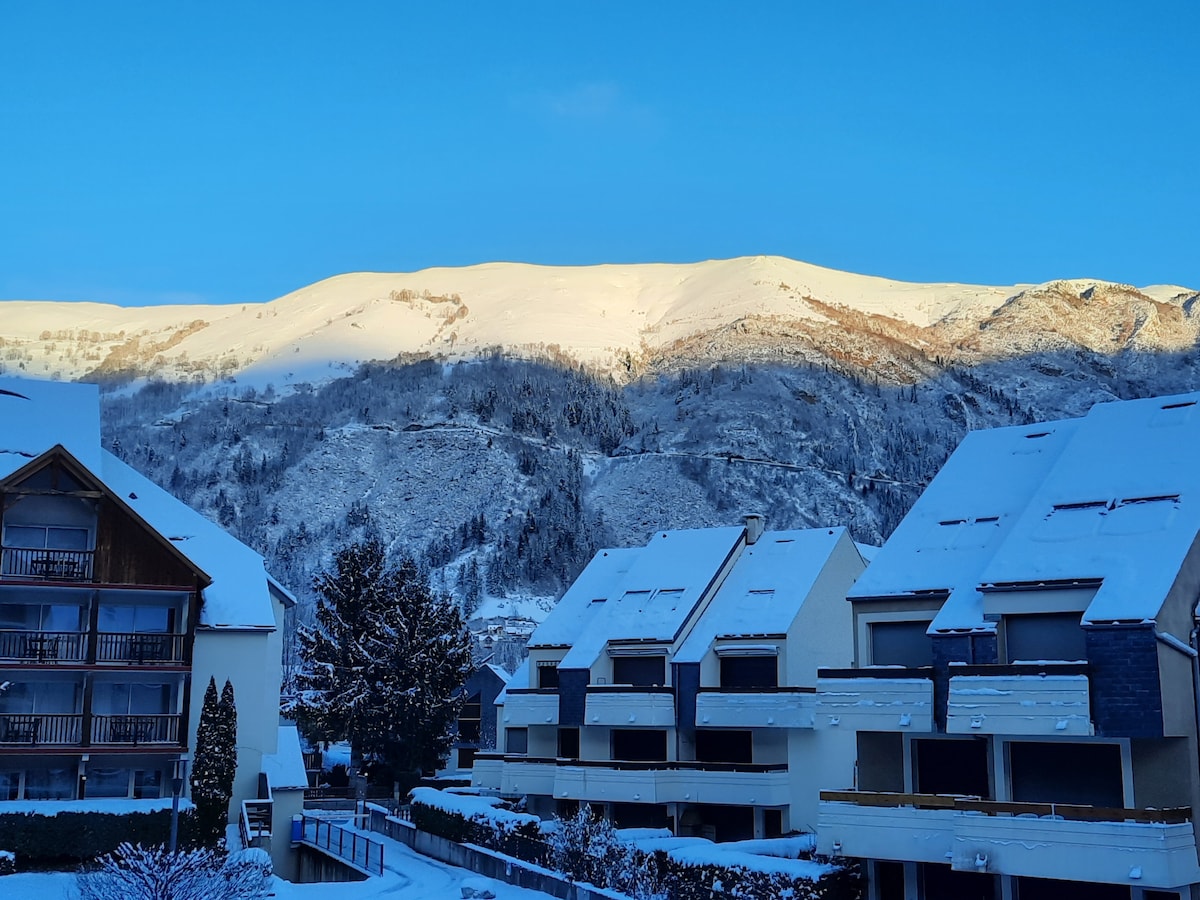 Snow-covered peaks rise majestically in the background, while modern apartment buildings are visible in the foreground. The serene wintry landscape is dominated by shades of white and light blue, creating a peaceful setting amidst nature.