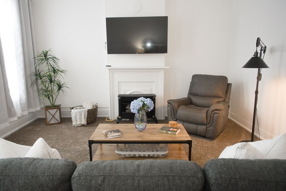 A comfortable living area features a gray sofa facing a wooden coffee table. A recliner is positioned adjacent to the table, with a floor lamp providing soft light. A television is mounted above a white fireplace, and a vase of blue flowers adds a touch of color.