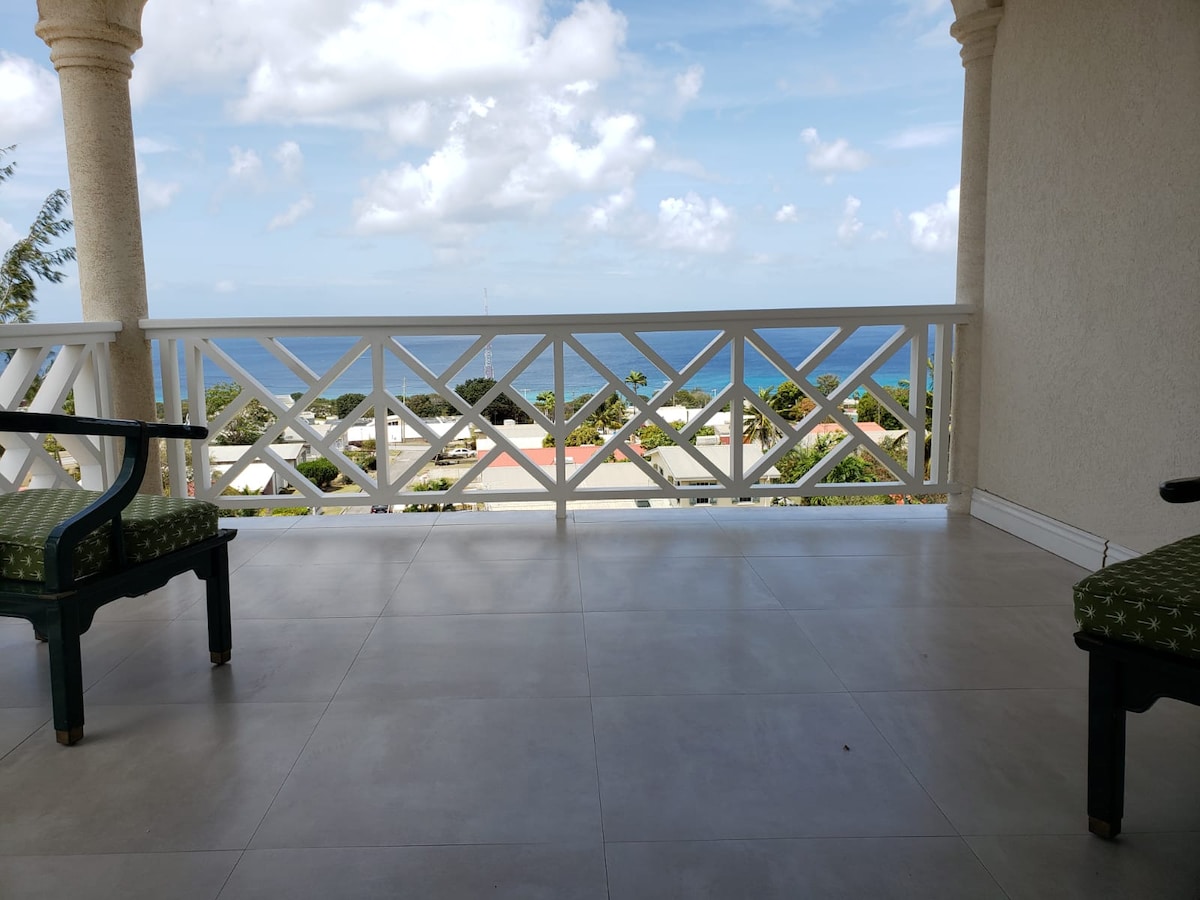 A spacious patio with a white railing overlooks the ocean and lush greenery. Two green chairs are positioned on the tiled flooring, providing a comfortable spot to enjoy sea views and nearby rooftops. The sky is bright and partly cloudy.