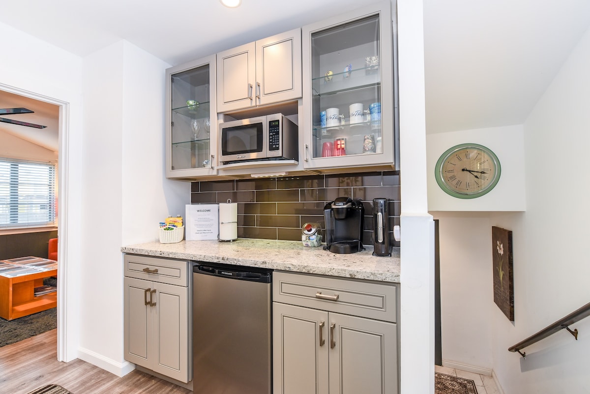 The coffee bar area features modern cabinetry with glass doors above showcasing dishware and mugs. A microwave and coffee maker are provided on the granite countertop, alongside a dishwasher. The setting is complemented by a contemporary clock on the wall, adding a functional element.