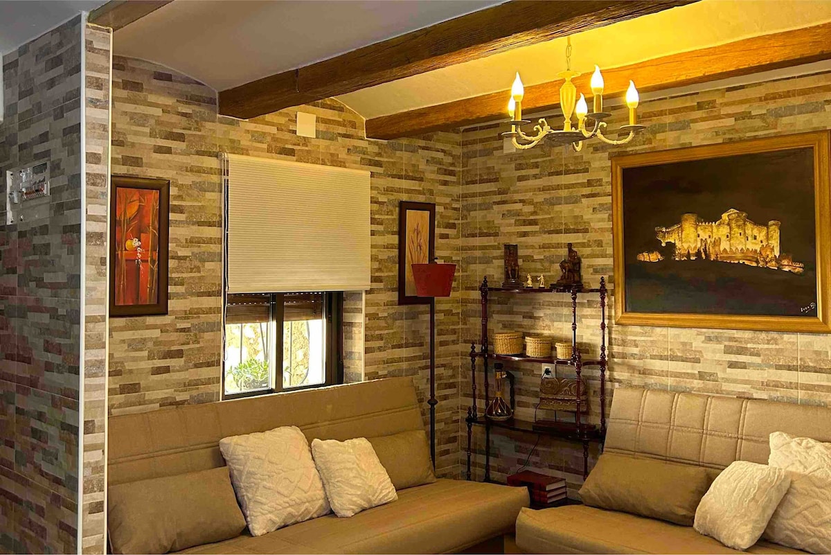 An inviting living area features textured stone walls and soft seating arrangements. A decorative chandelier provides warm lighting, while a wall-mounted artwork and shelves with decorative items add character to the space. Natural light filters through a window adorned with a simple blind.