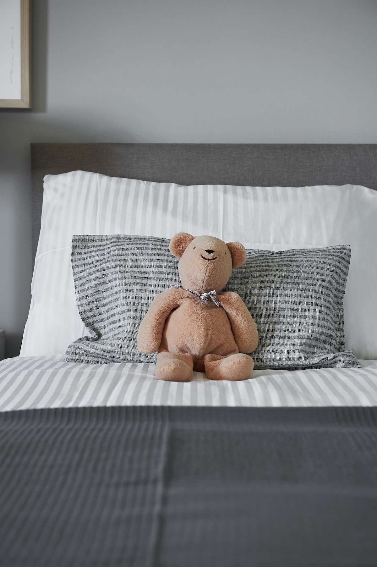 A cozy sleeping area features a spacious bed with a combination of striped and solid grey bedding. A soft teddy bear sits against the headboard, adding a touch of charm to the inviting setup.