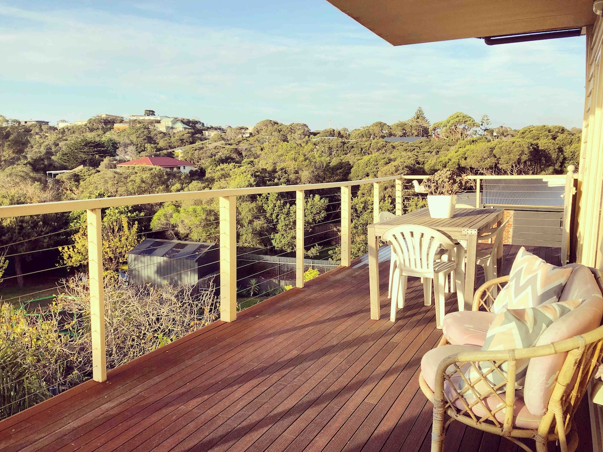 A spacious outdoor balcony is shown, featuring a wooden deck and comfortable seating. A small dining table is placed beside white plastic chairs, with views of lush greenery and distant rooftops visible across the landscape.