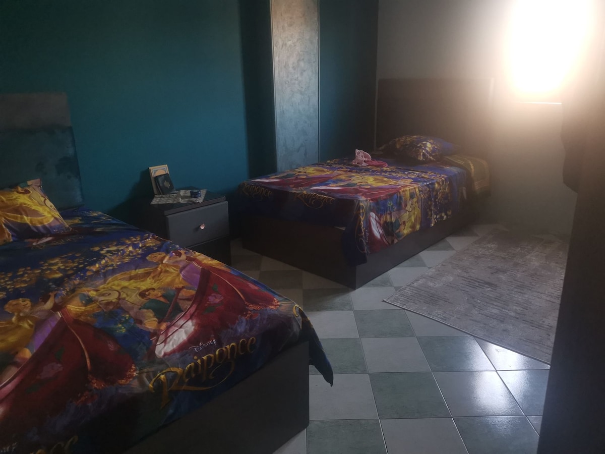 The room features two twin beds adorned with colorful, themed bedding. A bedside table stands next to one bed, and a light stream enters the space from a nearby window. The floor displays a glossy checkered pattern, complemented by a soft area rug.