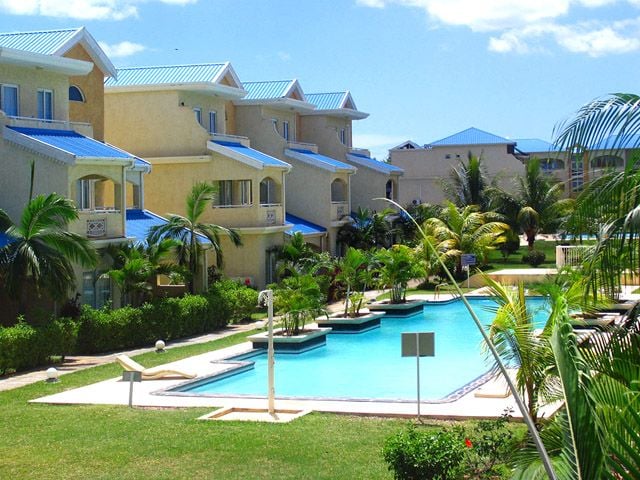The image showcases a residential complex featuring a central swimming pool, surrounded by well-maintained gardens adorned with palm trees and lush greenery. The two-story buildings exhibit bright blue roofs, contributing to the serene outdoor environment.