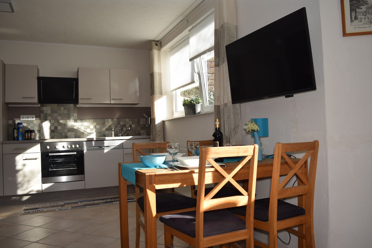 A modern kitchen area is visible, featuring grey cabinetry and stainless steel appliances. A wooden dining table is set for four, accompanied by a blue table runner. A flat-screen TV is mounted on the wall, and a small plant decorates the window sill.