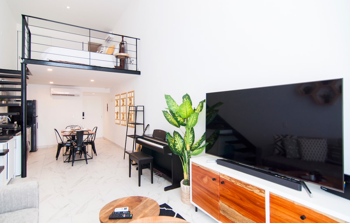 The spacious living area features a large 60-inch smart TV and a sleek wooden cabinet beneath it. A charming piano and a dining table with black chairs are visible. A tall green plant adds a touch of nature, while the lofted sleeping area is accessible by a modern staircase.