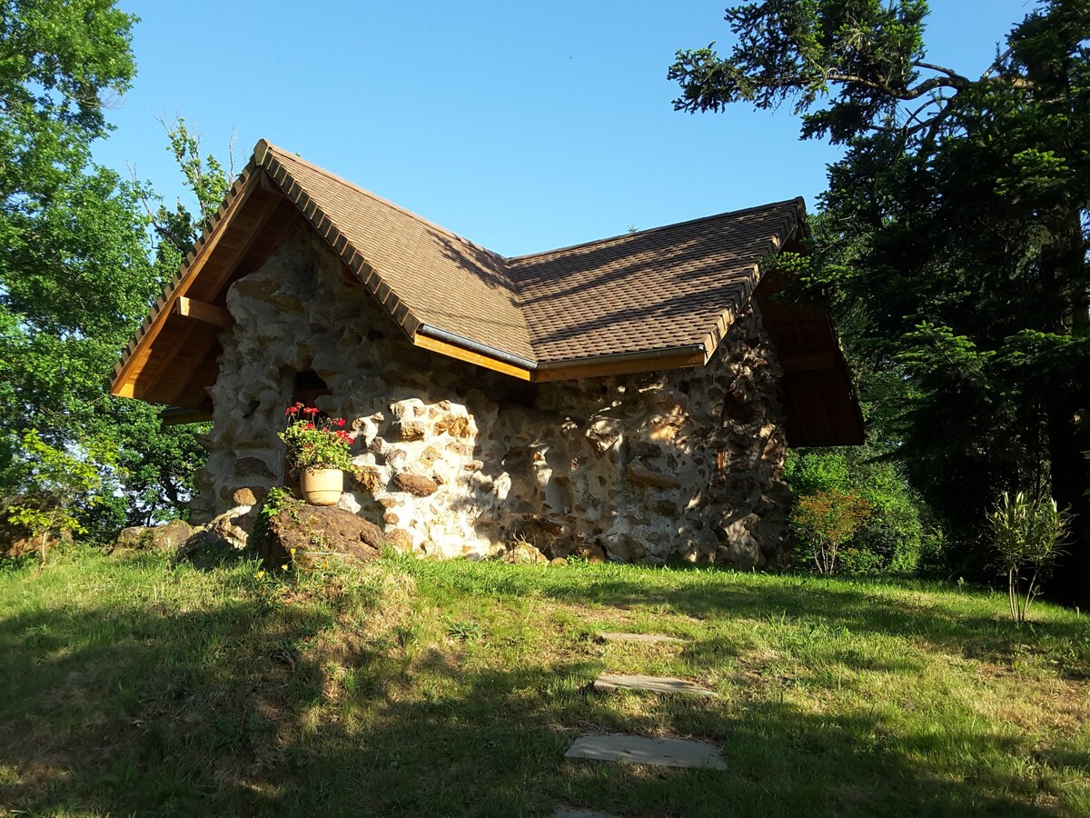 A charming stone cottage sits nestled among greenery, featuring a sloped roof and wooden eaves. A potted flower arrangement adds a touch of colour to the exterior, while stone pathways lead towards the entrance, inviting exploration of the natural surroundings.