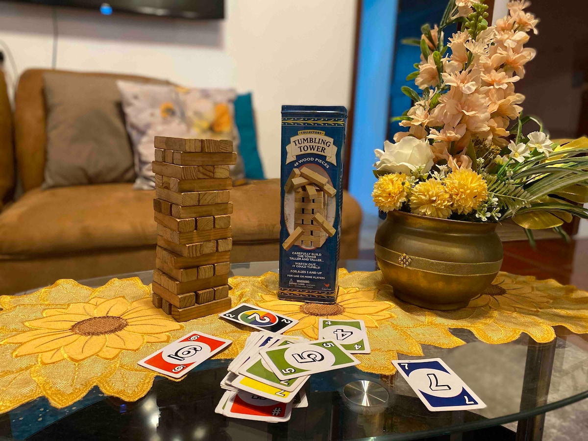 A coffee table is set with a carefully arranged vase of flowers, along with a colorful game of cards. A wooden stacking game, known as Tumbling Tower, is displayed next to the vibrant cards. The warm tones of the setting create a welcoming atmosphere.