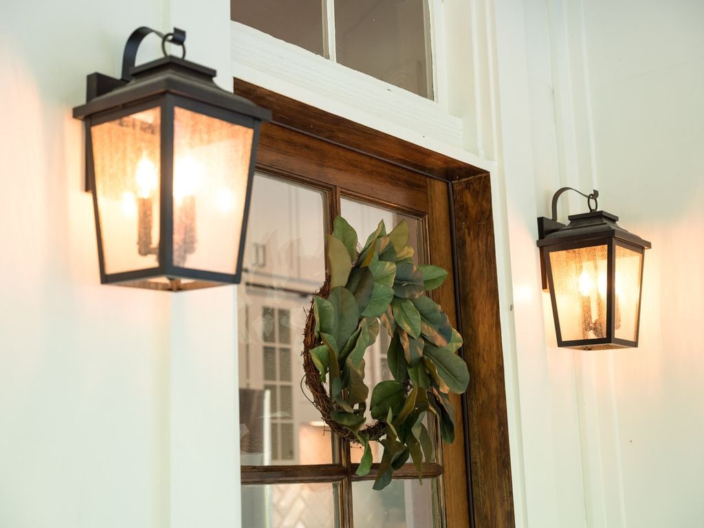 The entryway is framed by two black lantern-style light fixtures, casting a warm glow. A wooden door with glass panes leads inside, while a green wreath hangs prominently in the center, adding a touch of natural decor.