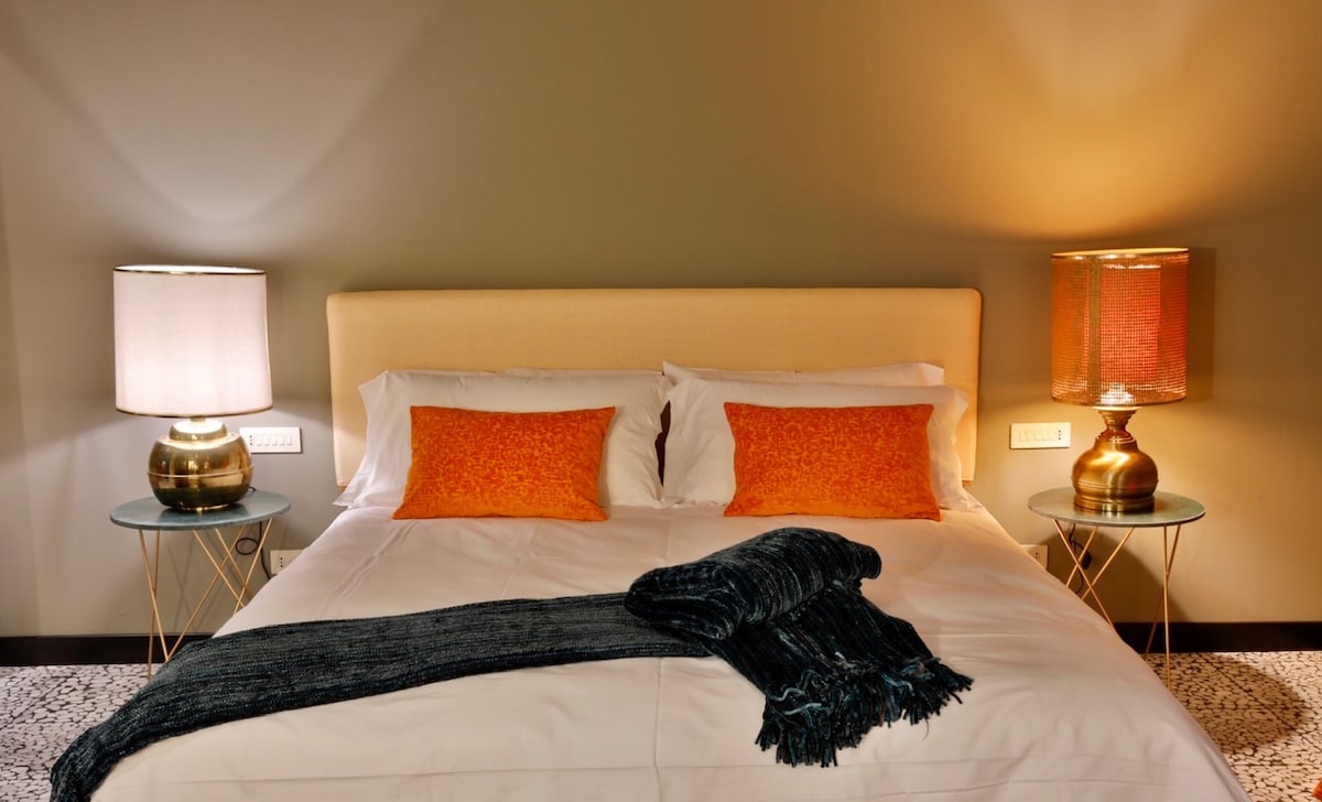 A well-appointed bedroom features a spacious bed with white linens and orange-accented pillows. Two bedside tables hold stylish lamps, casting a warm glow in the room. A textured blanket is draped over the bed, enhancing the inviting decor.
