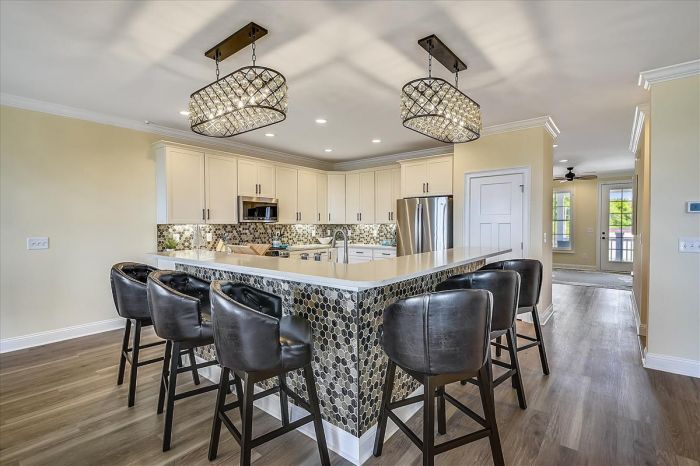 A modern kitchen features a large countertop with seating for several guests. Elegant pendant lights hang above, illuminating the space. The backsplash displays a mosaic pattern, complementing the cabinetry and appliances. Natural light filters in from nearby windows, creating a bright and functional area.