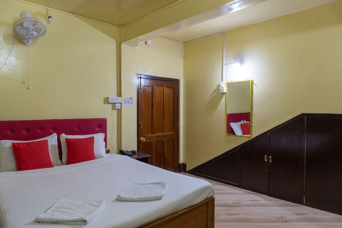 The room features a spacious bed with white linens and red accent pillows. A wooden door leads out, while a mirror hangs on the wall beside it. Light reflects off the walls painted in a soft yellow, contributing to a bright and clean environment.
