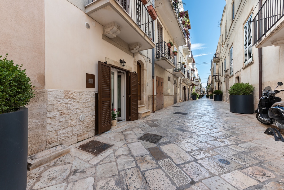 A charming street in a historic neighborhood is depicted, featuring warm stone buildings with wooden shutters. Potted plants enhance the entrance of the building. The cobblestone pathway leads to a quiet area, showcasing a motorcycle and planters lining the sides.