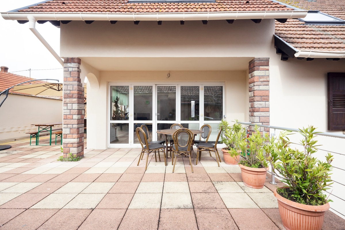 A spacious terrace features a round table surrounded by six chairs, complemented by potted plants. The terrace is paved and offers direct access through large windows, allowing for abundant natural light.