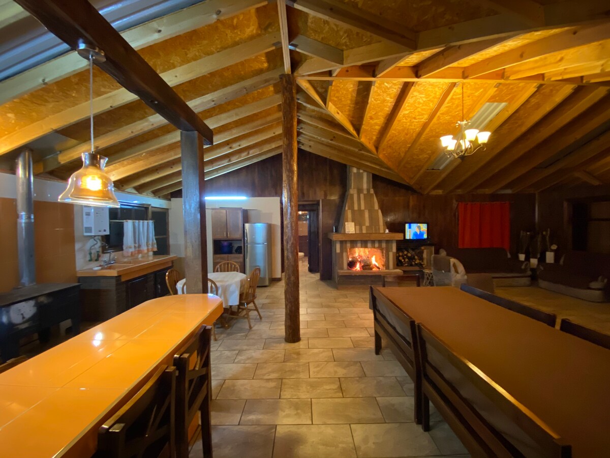 An open interior space is highlighted by a high, wooden beam ceiling and natural stone flooring. A cozy fireplace is positioned at one end, with a television nearby. A long dining table and several chairs accommodate group gatherings, while a kitchen area is visible in the background.