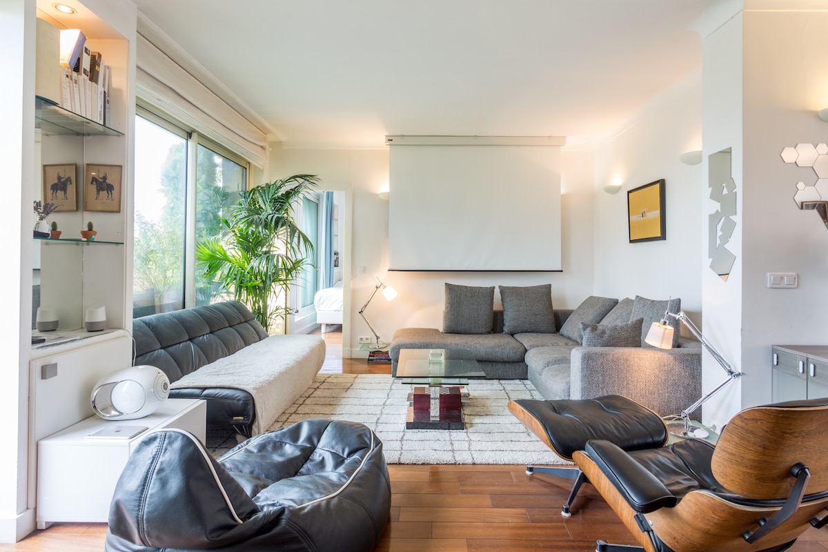 A spacious living area showcases modern furnishings, including a large sectional sofa, an inviting white sofa, and a stylish lounge chair. Natural light fills the space through expansive glass windows, highlighting a cozy rug and decorative plants. A projection screen is mounted on the wall.