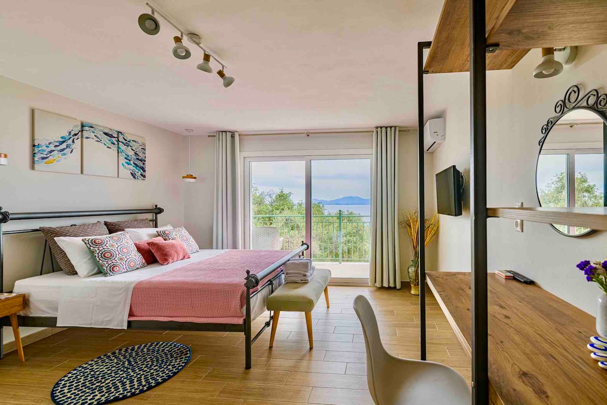 A bright and airy bedroom features a spacious bed with a pink throw blanket, complemented by decorative pillows. A wooden desk and chair are positioned next to a round mirror. Large French windows frame views of the sea and surrounding landscape, allowing natural light to fill the room.