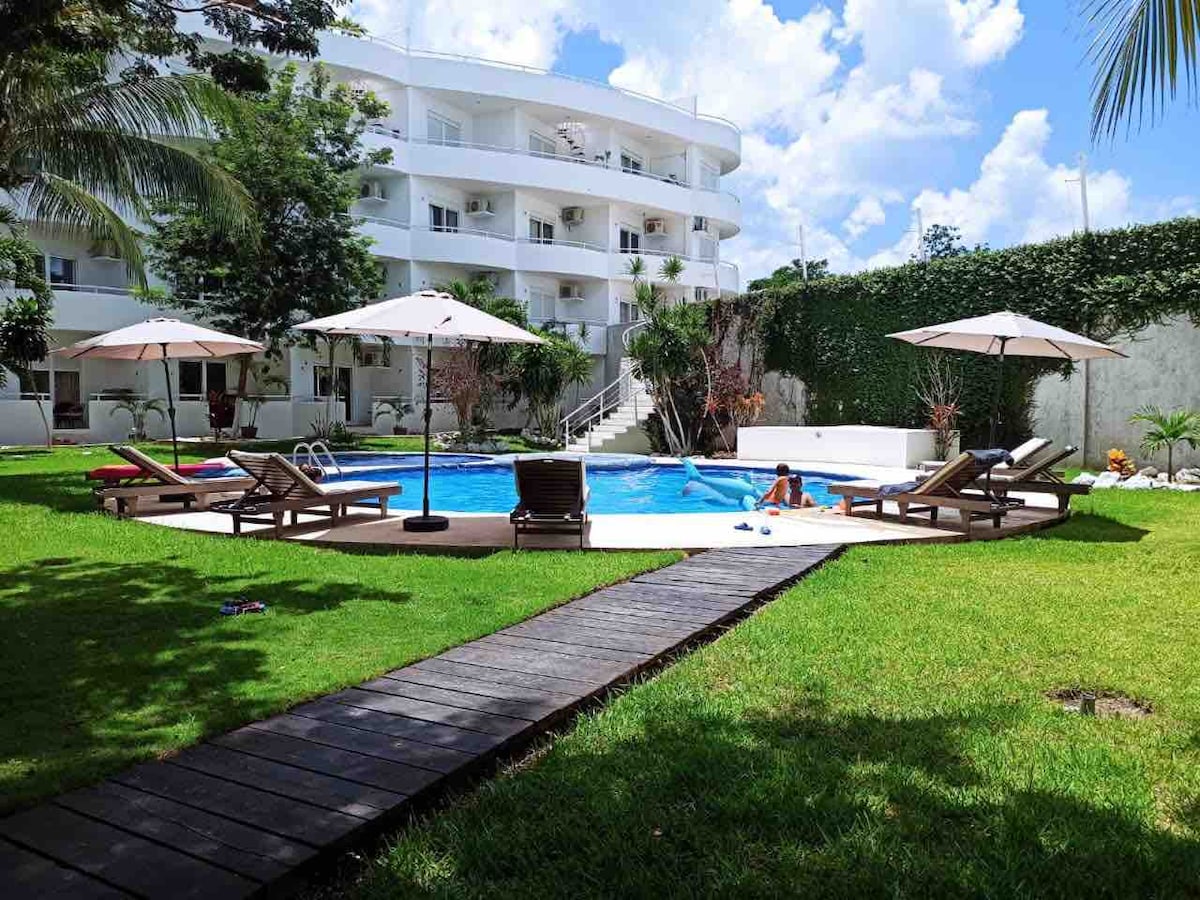 An inviting pool area is set within a lush green landscape. Sun loungers and umbrellas are positioned around the large circular pool. A wooden pathway leads through the well-maintained grass, connecting to the surrounding white-stucco building with multiple floors and balconies.