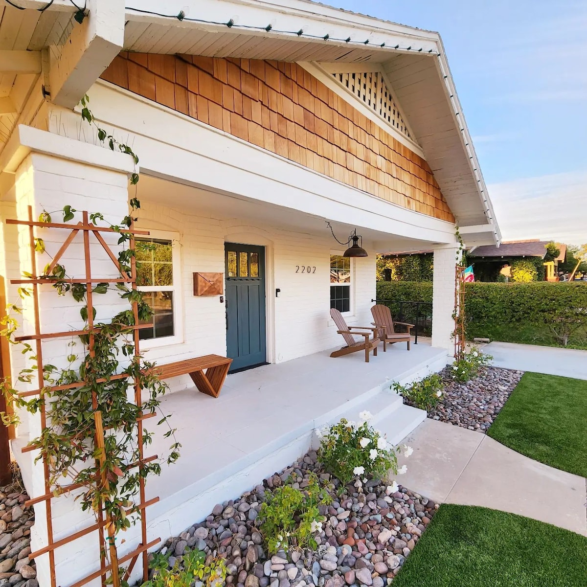 A charming front porch showcases a light blue door, flanked by two wooden Adirondack chairs. The area is adorned with climbing greenery and decorative stone, while fresh landscaping is visible in the foreground. Soft sunlight creates a welcoming ambiance.