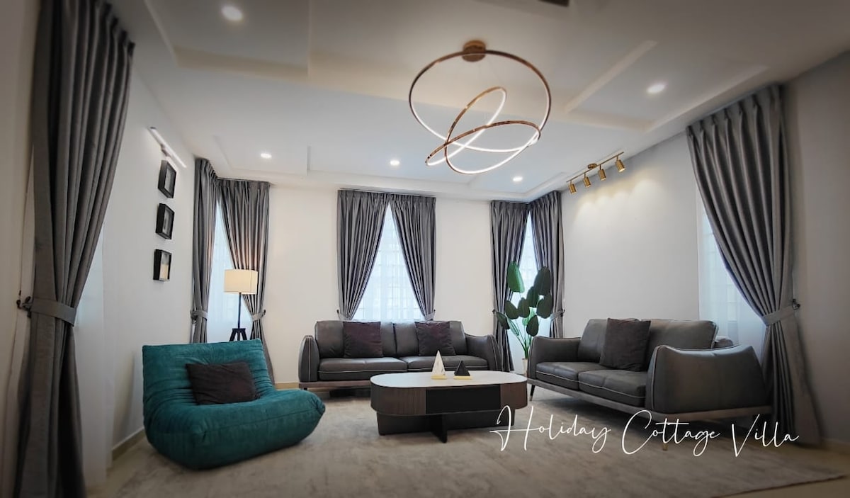 A modern living area is furnished with two gray sofas and a teal accent chair. A round coffee table is centered on a plush carpet, while large windows with gray drapes allow soft light to enter. A stylish light fixture hangs overhead.