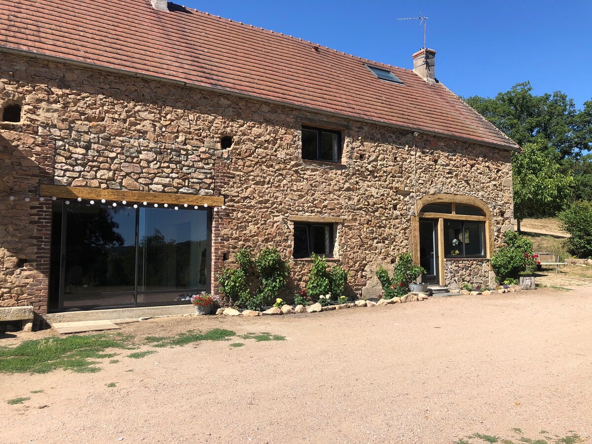 A rustic stone building is highlighted, featuring large glass doors that invite natural light inside. The entrance showcases a wooden arch, framed by greenery and colorful flowers. A gravel pathway leads towards the structure, set against a backdrop of trees and open land.