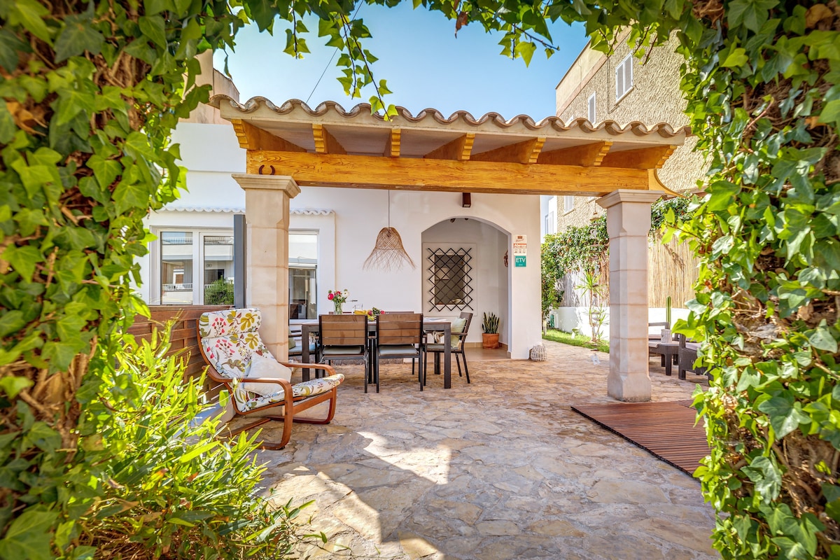 An outdoor dining area is framed by lush greenery, featuring a wooden table surrounded by six chairs. A cozy lounge chair with vibrant upholstery is positioned nearby. The covered terrace offers shade, enhancing the inviting atmosphere for gatherings.