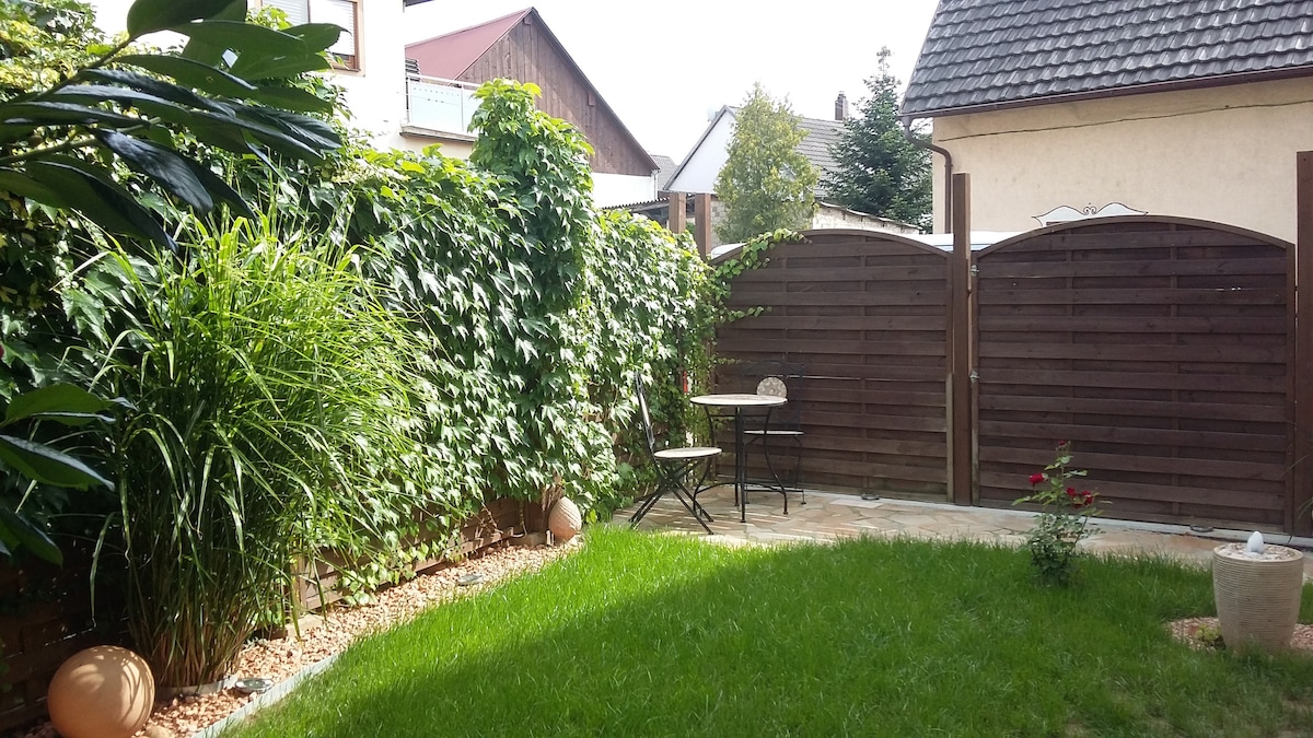 A serene garden area is surrounded by lush greenery and climbing plants. A small, round table is paired with two chairs, set against the wooden fence that provides privacy. Soft grass covers the ground, with decorative stones and a small flowerbed adding to the peaceful setting.