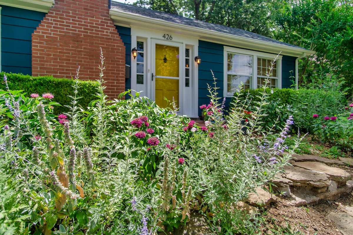 The charming entrance features a bright yellow door framed by tasteful landscaping. Lush greenery and colorful flowers, including pink and purple blooms, create an inviting atmosphere. The brick exterior contrasts with the blue siding, enhancing the home's vintage appeal.