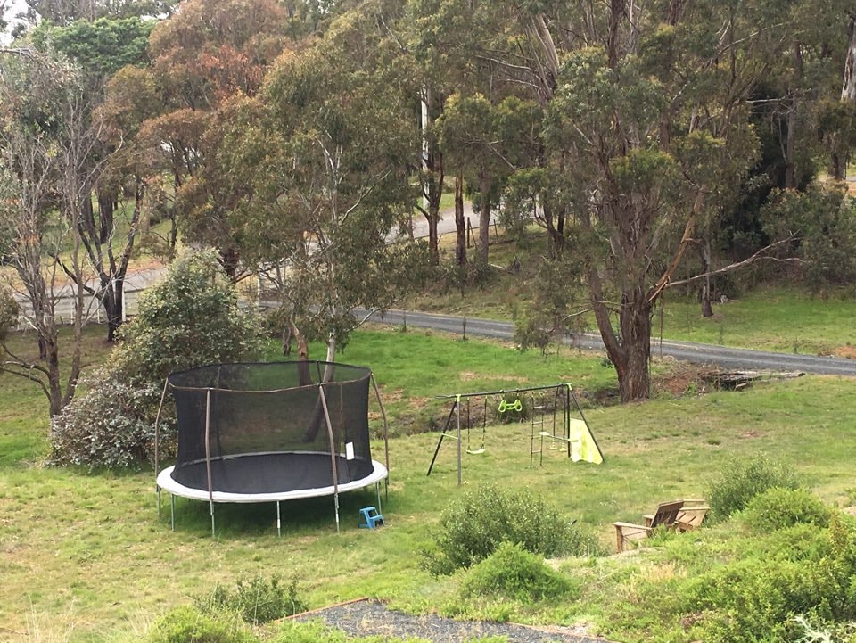 An expansive outdoor area is visible, featuring a large trampoline surrounded by lush greenery. A playground set is present, including swings and a small slide, positioned on a grassy space. A wooden bench seat offers a comfortable spot for relaxation nearby.