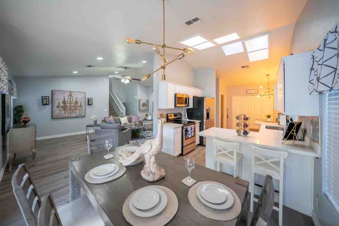 An open-concept living space showcases a modern kitchen and dining area. The dining table is set with plates and glassware, while stylish lighting fixtures hang above. A comfortable seating area with a sofa and decorative elements can be seen in the background, creating a welcoming atmosphere.