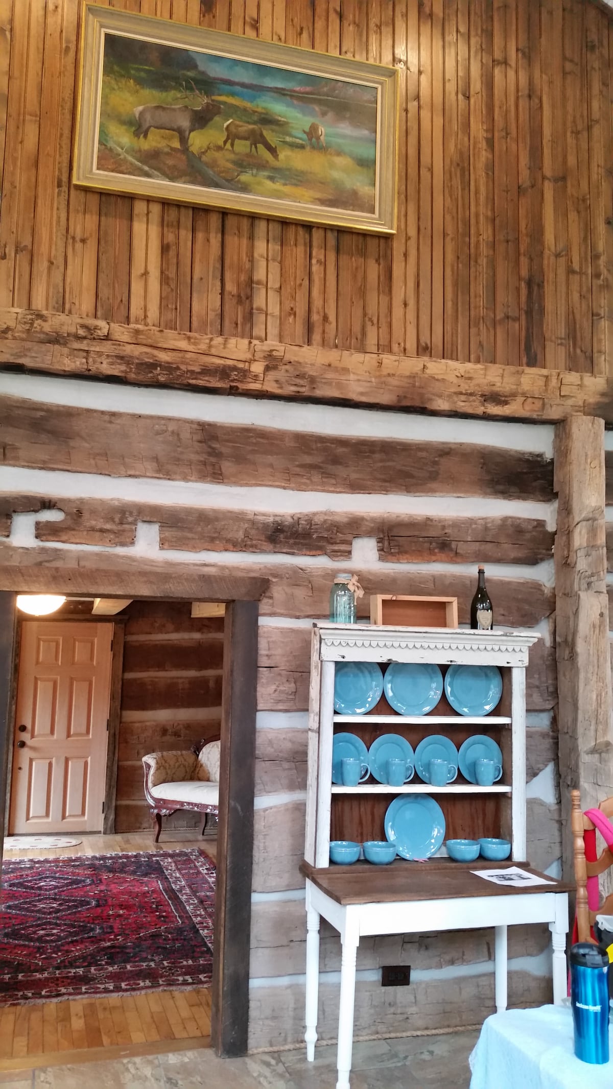 The interior features rustic wooden walls, emphasizing the charm of the restored log home. A collection of light blue dishware is displayed on a white wooden shelf. A framed painting hangs above, depicting a serene landscape. A patterned area rug adds warmth to the space.