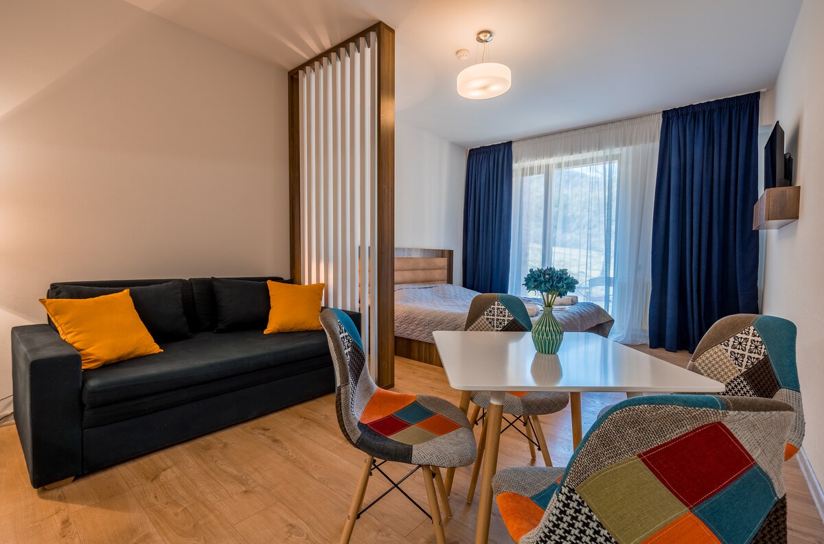 A bright living space features a cozy sofa with colorful cushions, a dining table with patterned chairs, and a spacious bed behind a wooden divider. Large windows provide natural light and showcase views of the outdoors, framed by deep blue curtains.