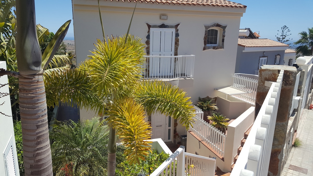 The exterior of a villa is presented, featuring a two-level structure with a white façade and terracotta roof. Lush palm trees and greenery are visible surrounding the stairs leading to the entrance. A sunny sky enhances the tranquil atmosphere of the setting.