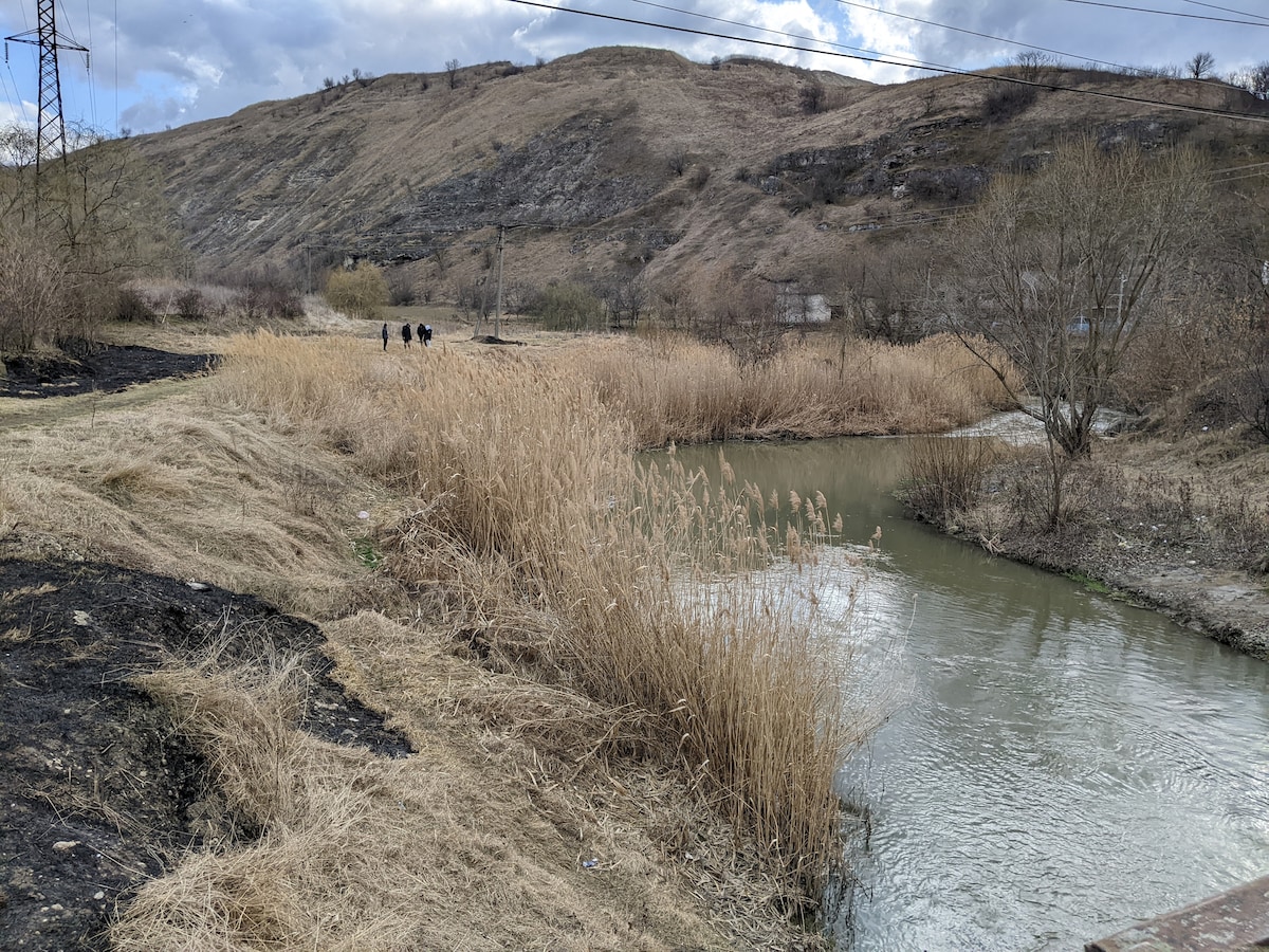 A calm waterway flows through a natural landscape adorned with tall grasses along the banks. Gentle hills are visible in the background, featuring sparse vegetation under a cloudy sky. A few individuals traverse the pathway on the opposite side of the water.