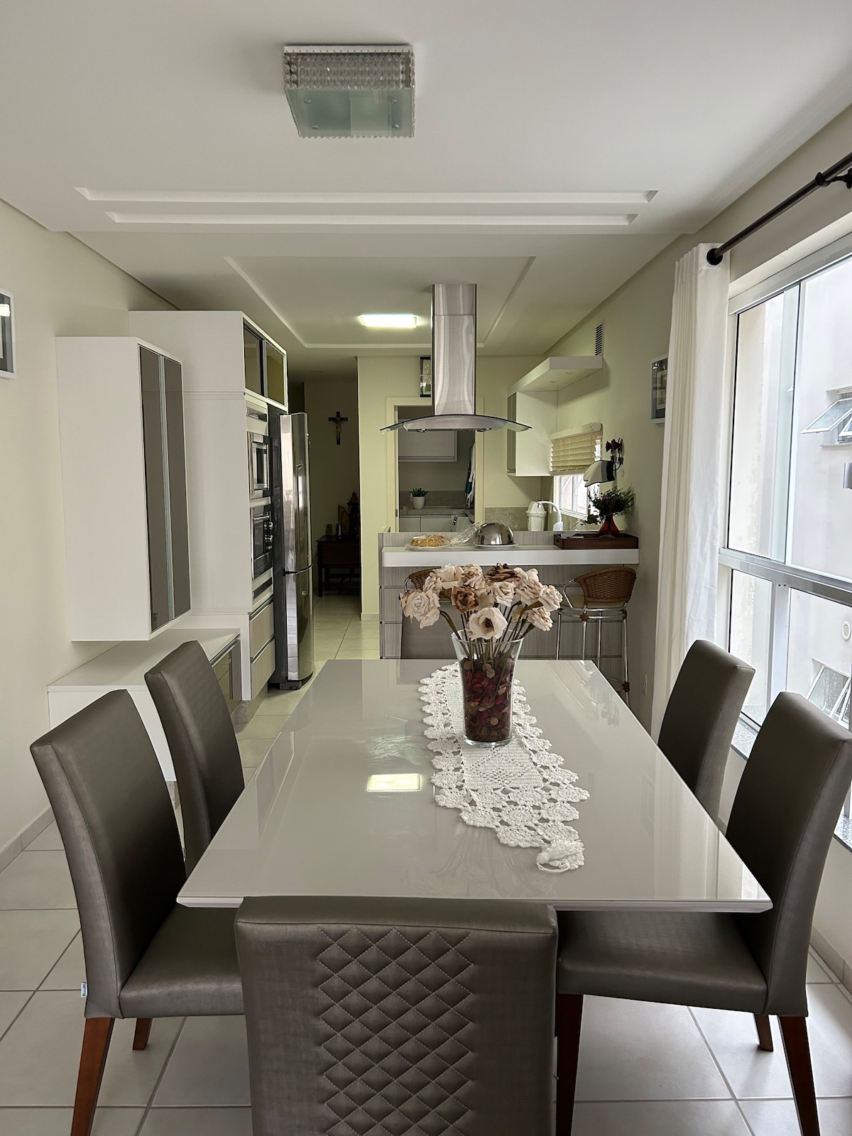 A modern kitchen features a polished dining table surrounded by six upholstered chairs. A decorative table runner with flowers adds a touch of elegance. The kitchen is well-equipped with sleek cabinetry and stainless steel appliances, while natural light enters through the large window.