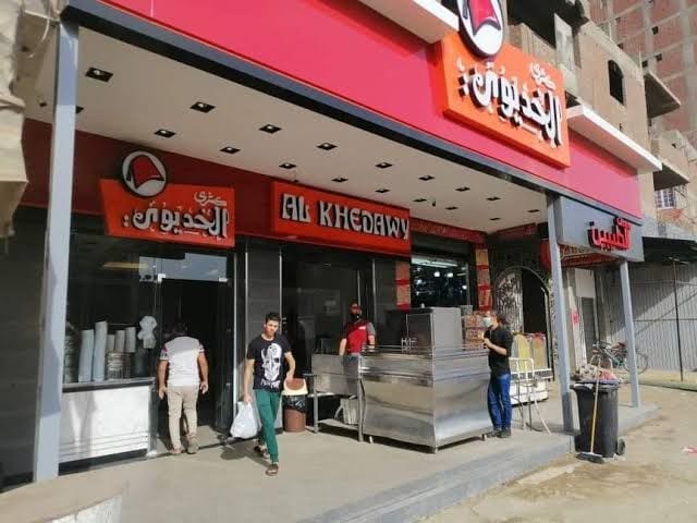 A vibrant restaurant exterior features a modern design with large signage displaying the name 'Al Khedawy.' Customers are seen interacting at the entrance, which is lined with large windows allowing for views of the bustling atmosphere outside. Outdoor seating is partially visible.