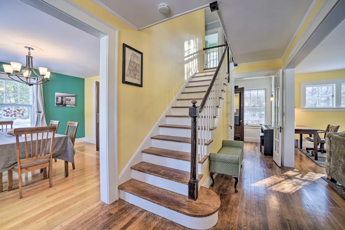 A well-lit entrance features a wooden staircase leading to the upper level, complemented by warm yellow walls. To the left, a dining area showcases a large table with wooden chairs, and to the right, a cozy living space is visible through an open doorway.