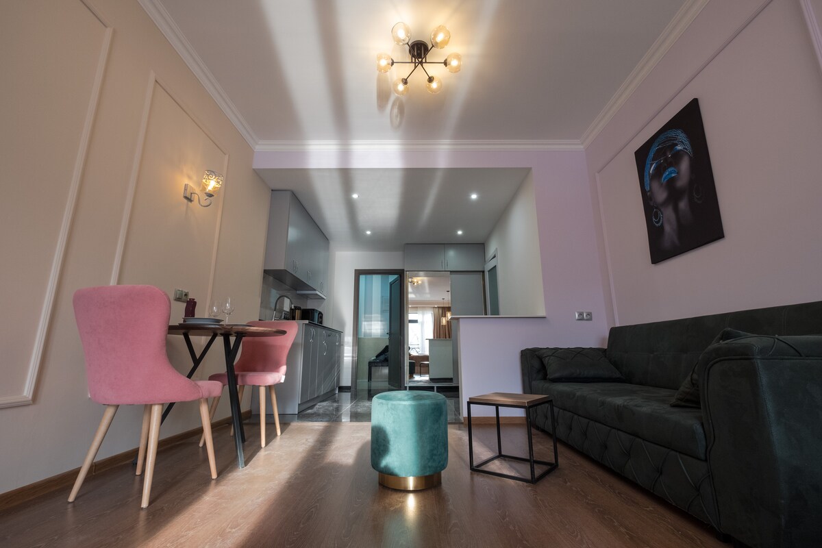 An open living area features a dark green sofa and a small wooden table with two stylish pink chairs. A round green ottoman sits in the center, and modern lighting fixtures hang overhead. The kitchenette is visible in the background, enhancing the space's functionality.