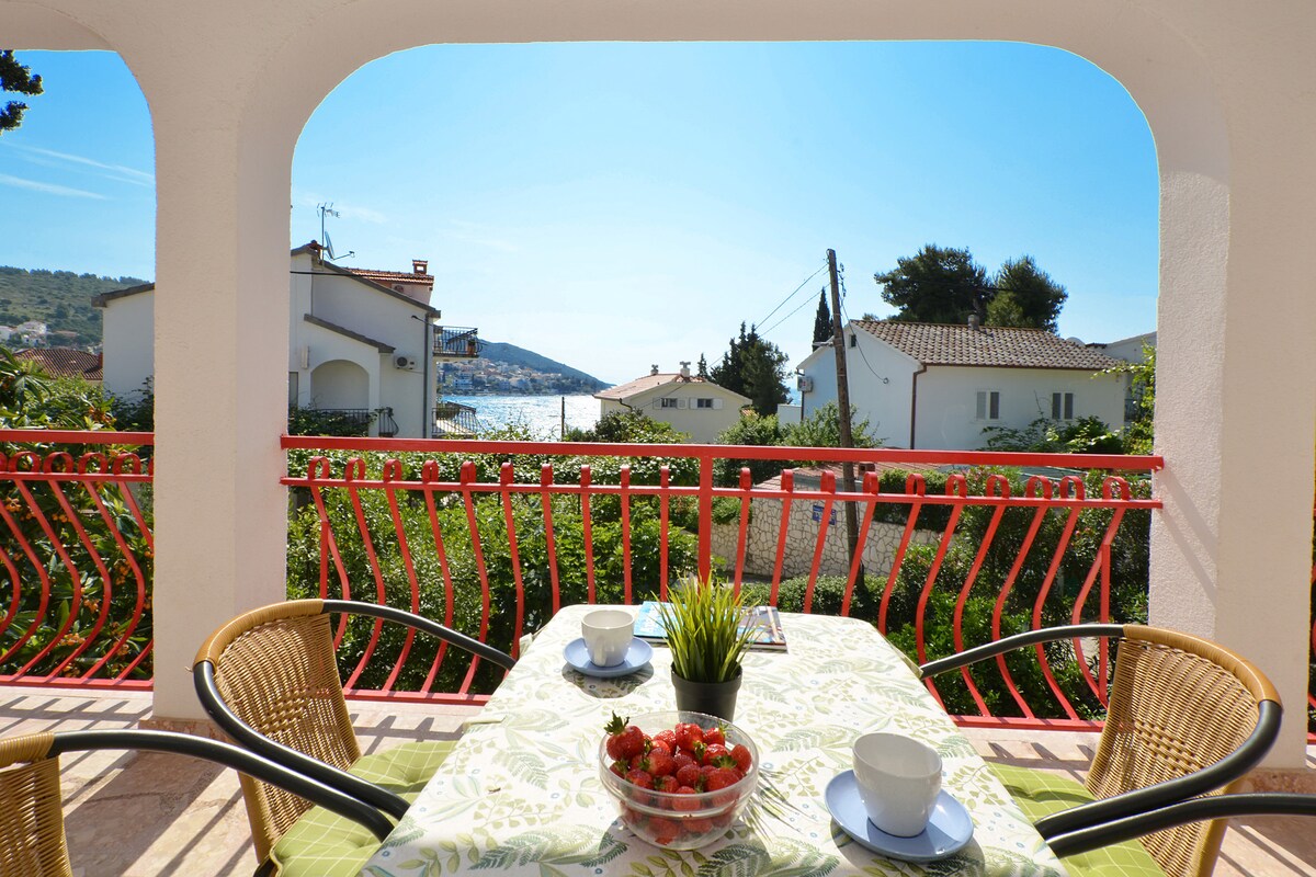 A spacious terrace features a table set for two, adorned with a green tablecloth and a bowl of fresh strawberries. The view opens up to the sea and surrounding landscapes, framed by an archway with a vibrant red railing.
