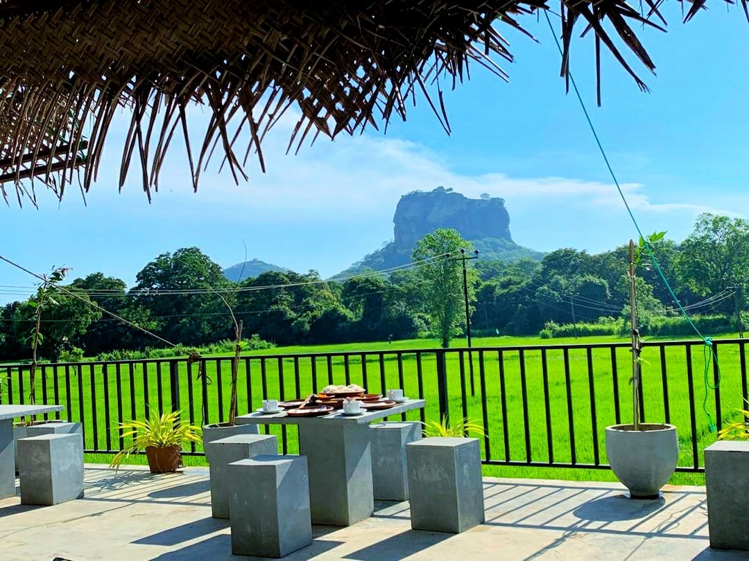 An outdoor dining area features concrete tables and stools, with a backdrop of lush green fields. A prominent rock formation rises in the distance under a clear blue sky, framed by trees and power lines.