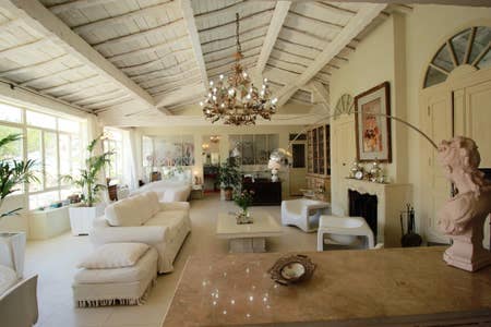 A spacious living area features high ceilings with exposed beams and large windows allowing natural light. The room contains a mix of seating arrangements, including plush sofas and stylish chairs. Decorative plants and a chandelier enhance the elegant atmosphere.
