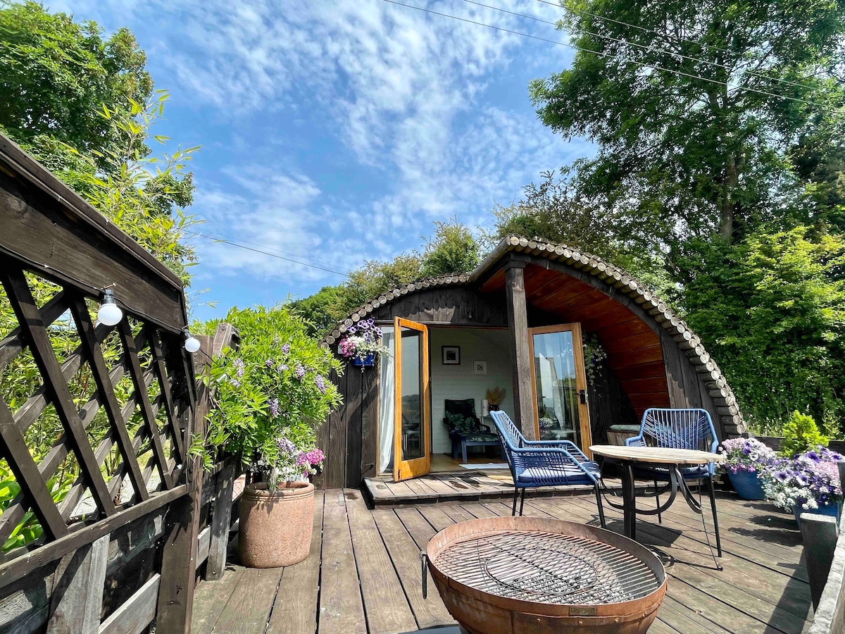 A wooden deck area features a round cabin surrounded by greenery. Outdoor seating is provided with a small table and two blue chairs. Potted plants add color, and a fire pit is visible in the foreground under a clear blue sky.