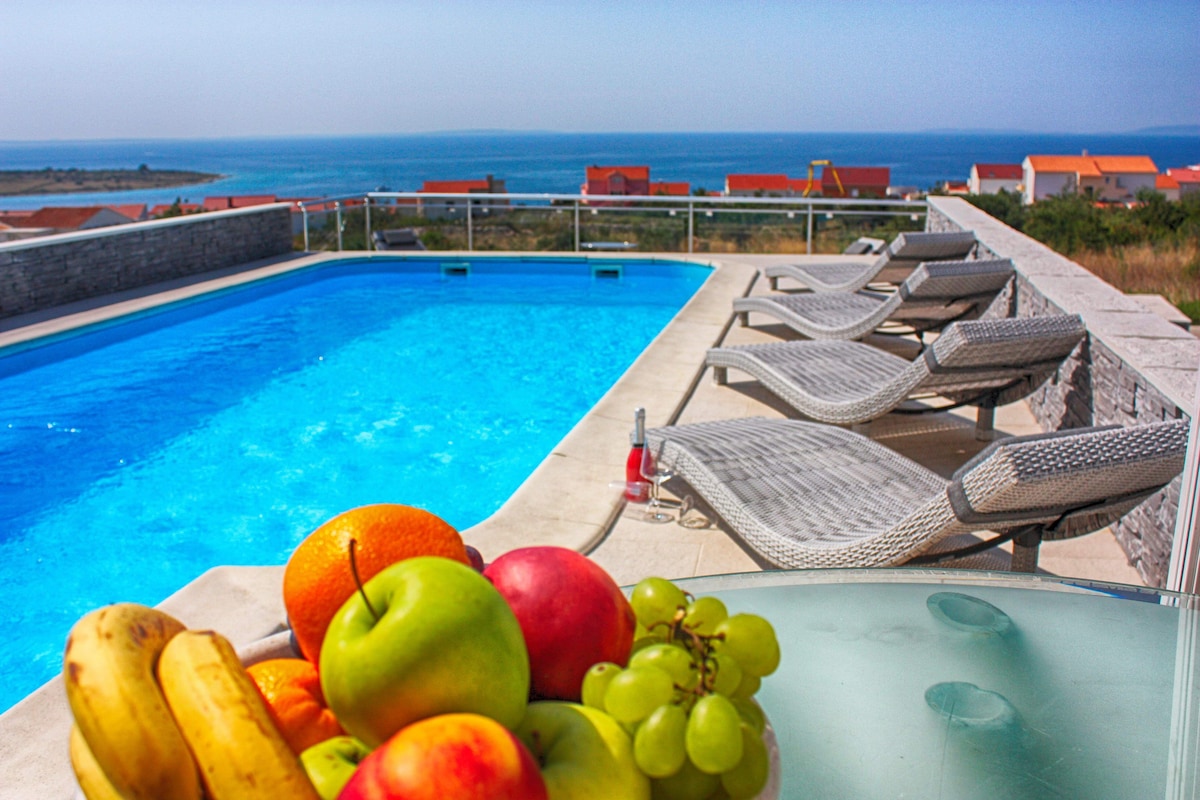 A private pool is surrounded by lounge chairs, offering clear views of the sea. A bowl of various fruits, including bananas, apples, and grapes, is prominently positioned in the foreground. The tranquil blue water reflects the sky, enhancing the serene atmosphere.