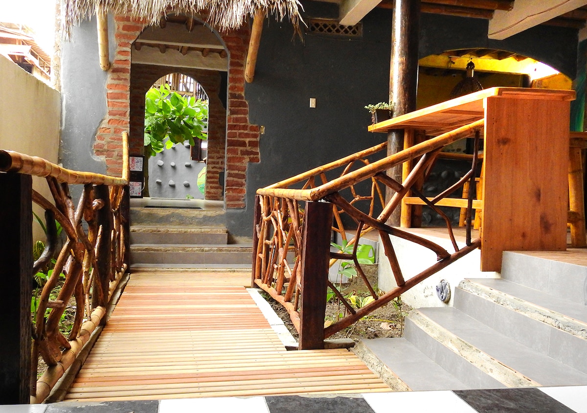 A wooden pathway leads through a charming entryway, framed by bamboo railings with a natural design. Stone steps descend into a lush green space, with rustic brick walls visible in the background, creating an inviting transition between indoor and outdoor areas.