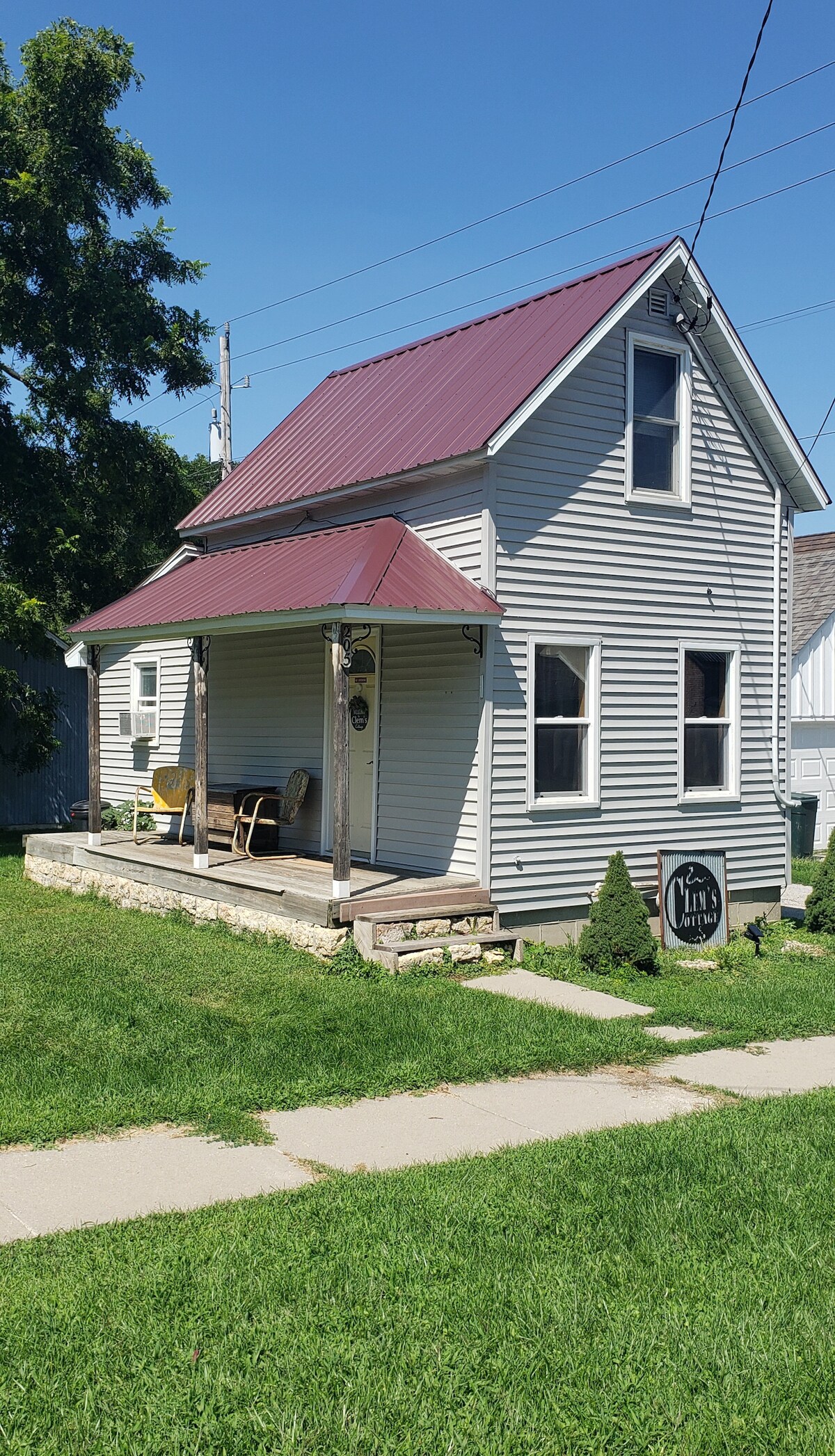 The cottage presents a charming exterior with light grey siding and a maroon roof. A small porch with seating is visible, surrounded by a well-maintained lawn and landscaping. The entrance features a welcoming sign, indicating the cottage's name.